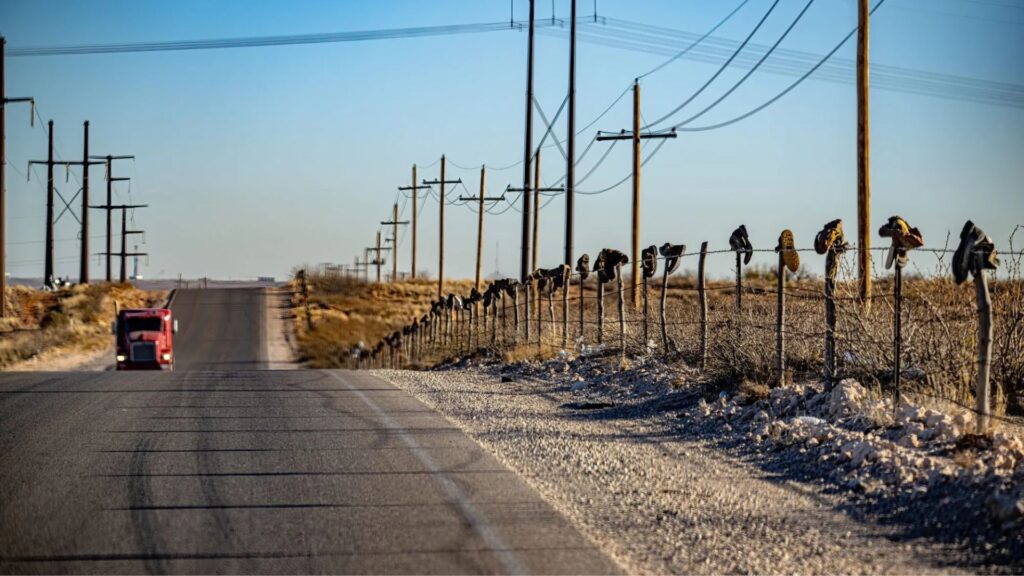A rural road with a red vehicle driving away; shoes are hanging on a wire fence running alongside utility poles under a clear blue sky.