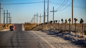 A rural road with a red vehicle driving away; shoes are hanging on a wire fence running alongside utility poles under a clear blue sky.