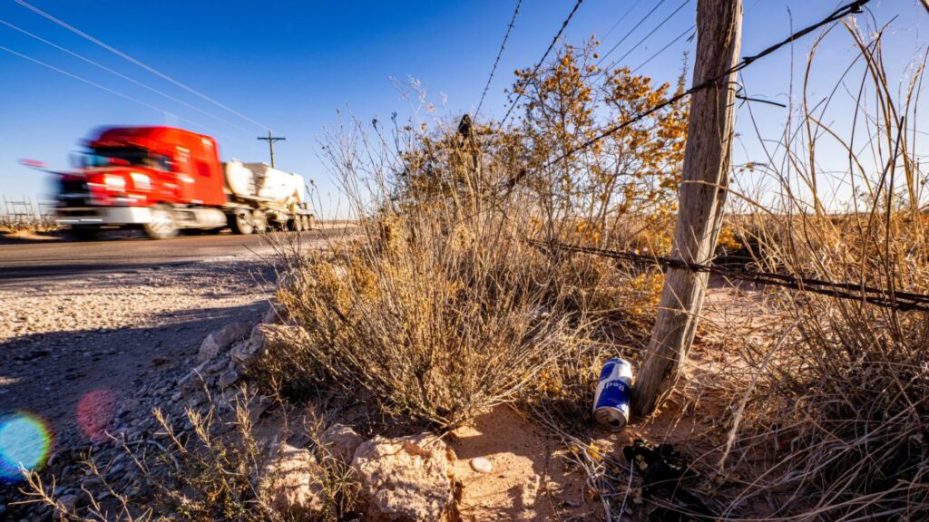 A red semi-truck drives down a rural road past a barbed wire fence, dry grass, and a discarded blue-and-white can in the foreground under a clear blue sky.