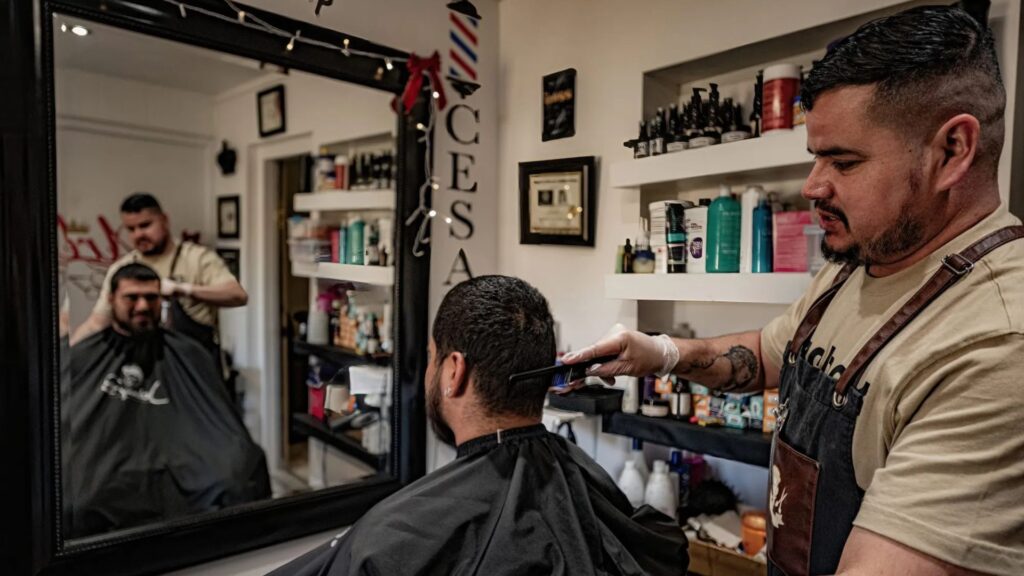A barber trims a customer's hair in a well-equipped barbershop, with both visible in the large mirror in front of them.