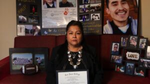 A woman sits on a red couch holding a San Juan College certificate, surrounded by framed photos and posters featuring a young man and family pictures.