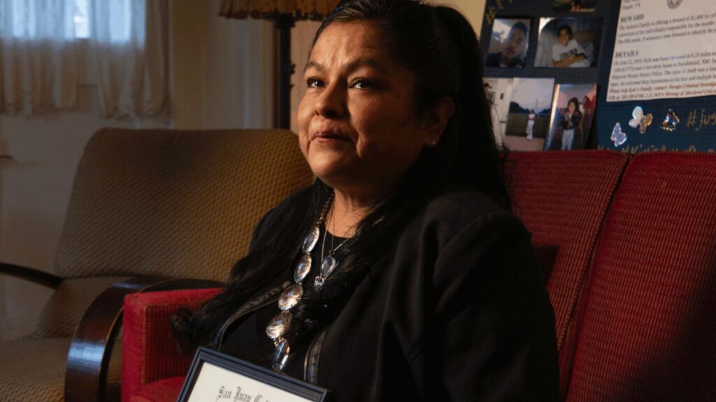 A woman with long dark hair sits on a red chair indoors, holding a framed document, with photos and memorabilia displayed in the background.