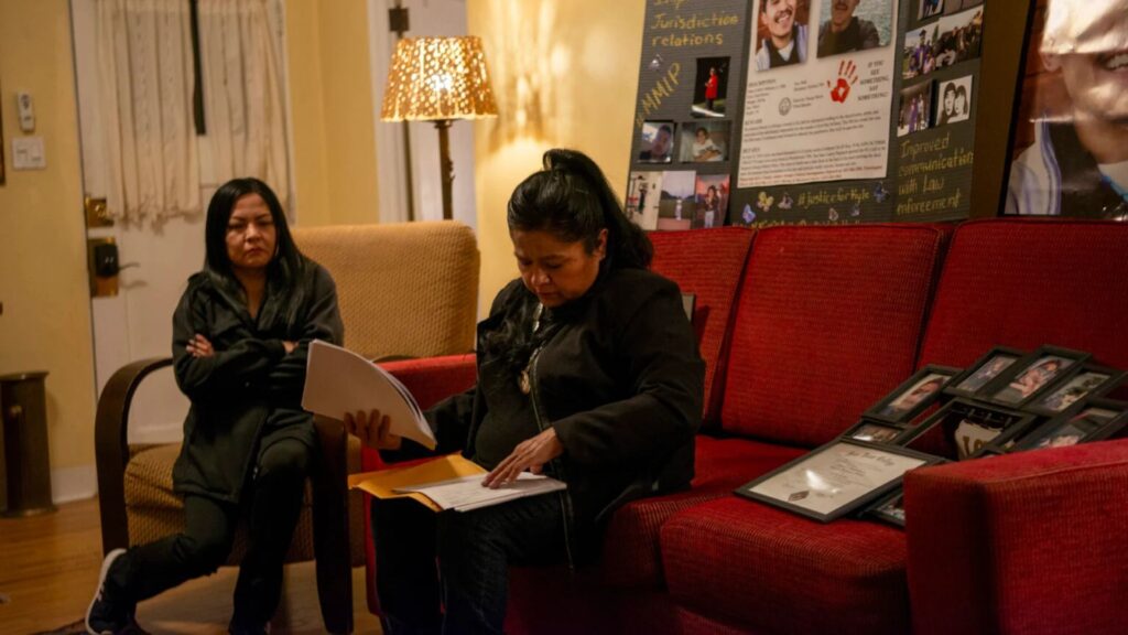 Two women sit in a living room; one looks at documents on a red couch, while the other sits nearby. Framed photos and a display board are visible in the background.