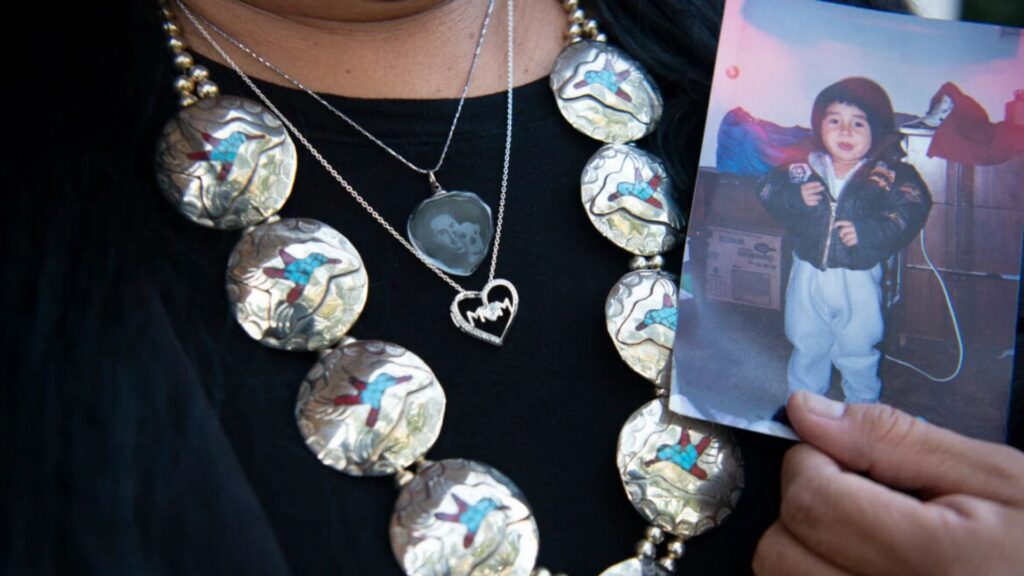 A person wearing silver jewelry holds up a photo of a young child in a jacket and boots.