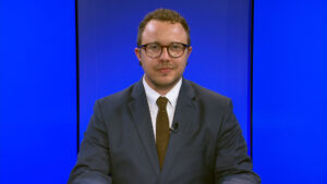 A man wearing glasses and a suit sits in front of a blue background, looking directly at the camera.