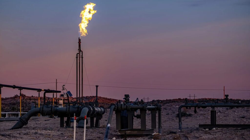 Natural gas is flared from a tall pipe at an industrial facility in a barren landscape during dusk, with various pipes and equipment visible in the foreground.