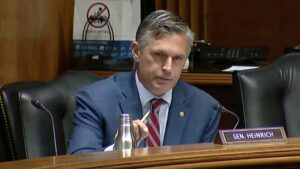 A man in a suit and tie sits at a desk with a nameplate reading "Sen. Heinrich," holding a pen during a meeting or hearing.