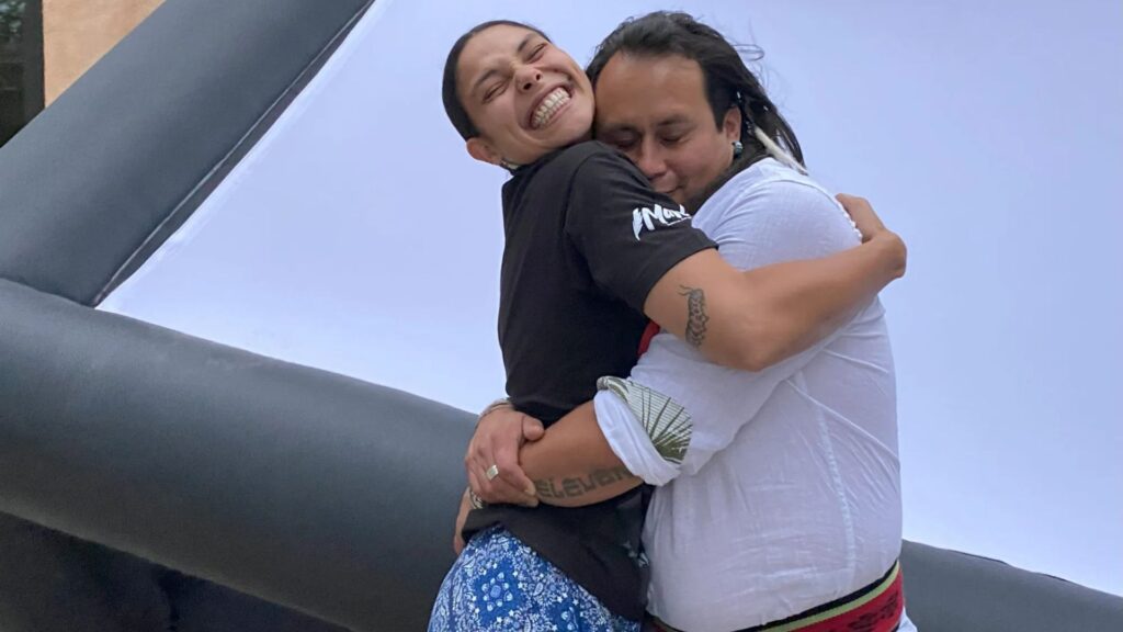 Two people hug each other tightly and smile, standing in front of a large inflatable black and white structure outdoors.