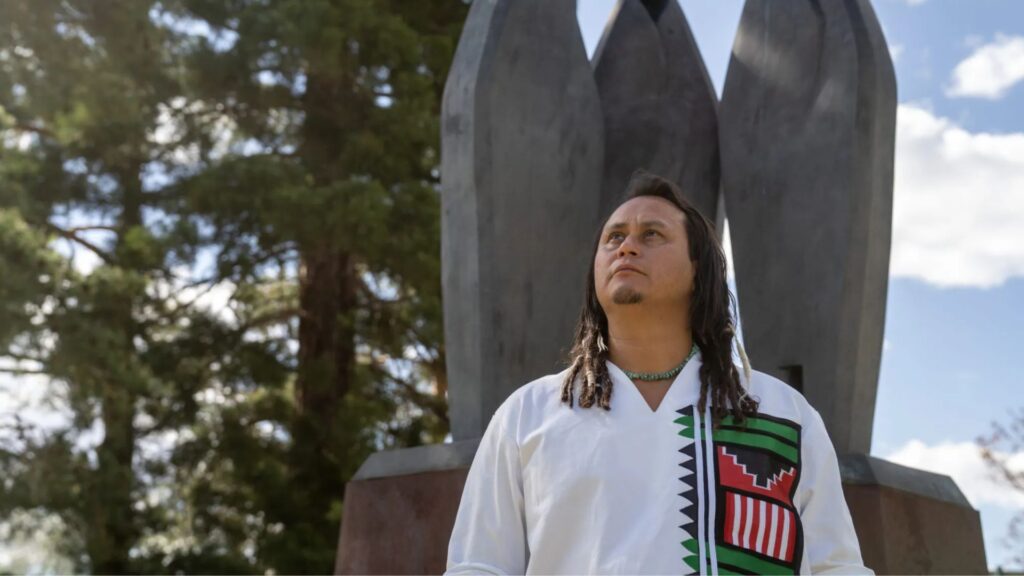A person with long hair and a patterned shirt stands in front of a tall, abstract outdoor sculpture with trees and sky in the background.