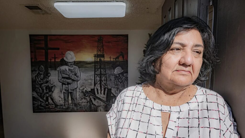 A woman with short dark hair stands indoors near a window; behind her is a painting of oil field workers with a sunset sky.