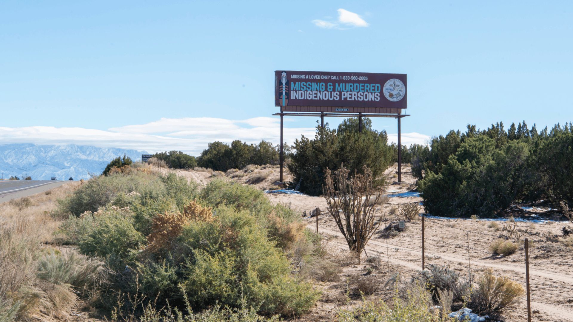 A roadside billboard reads "Missing & Murdered Indigenous Persons" with contact information, set in a desert landscape with bushes and distant mountains.