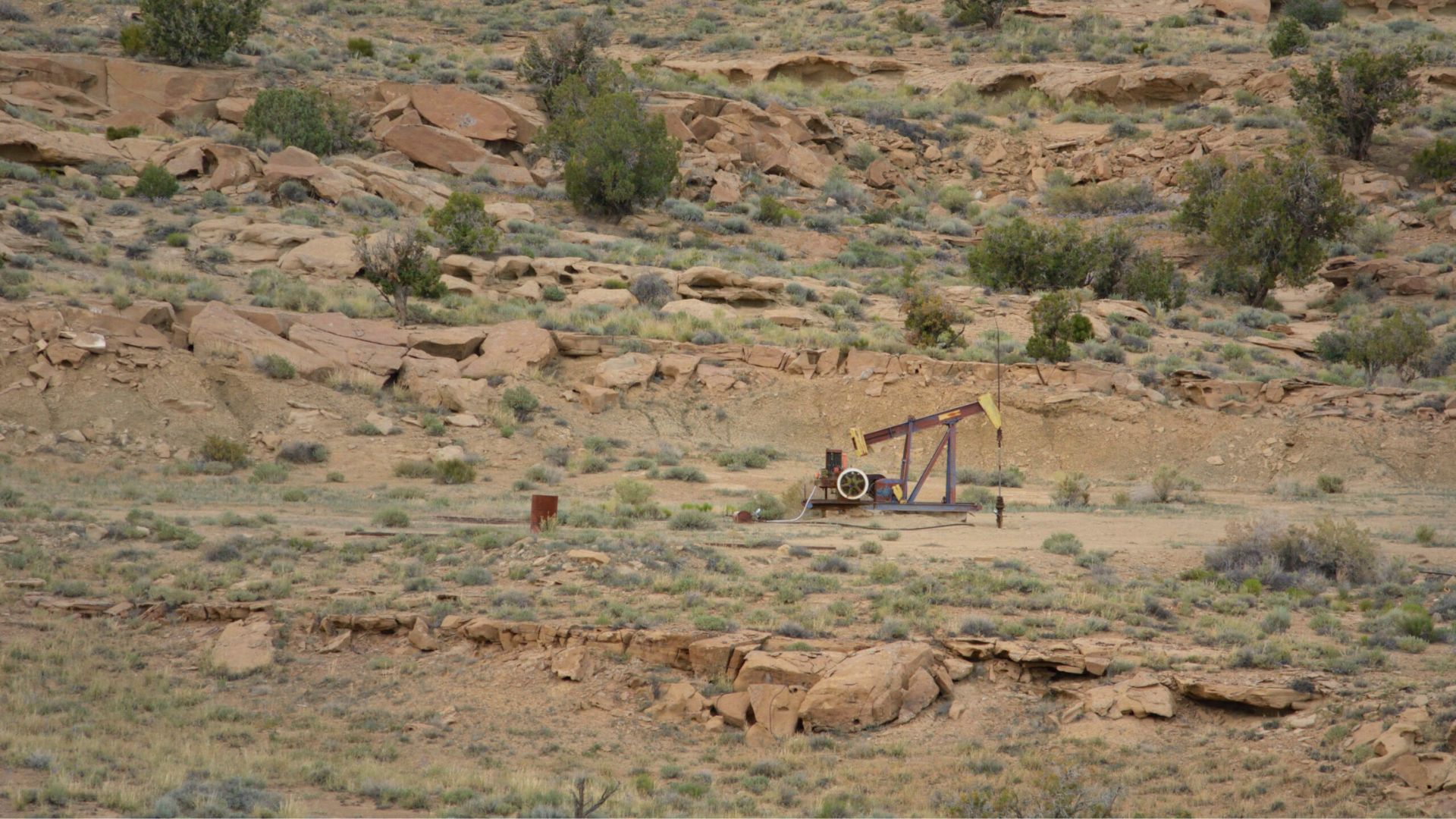 A single oil pumpjack operates in a dry, rocky landscape with sparse vegetation and scattered shrubs.