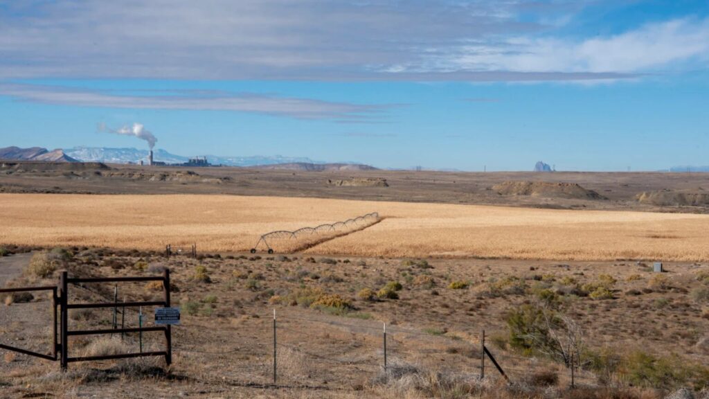 A fenced, dry landscape with a circular irrigation system crosses a golden crop field; smoke stacks and mesas are visible in the distant background under a partly cloudy sky.