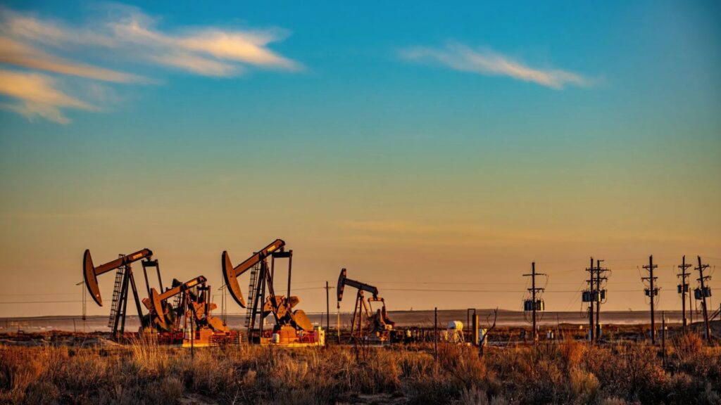 Several oil pumpjacks operate in a dry, grassy landscape at sunset, with power lines and a blue sky with clouds in the background.