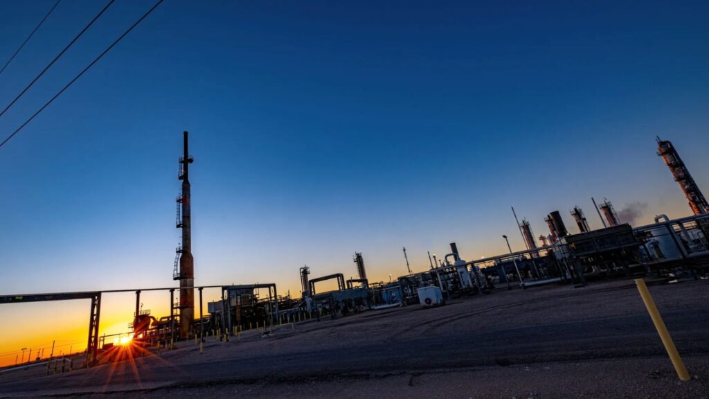 Industrial refinery structures silhouetted against a clear sky at sunset, with various towers, pipes, and equipment visible across the scene.