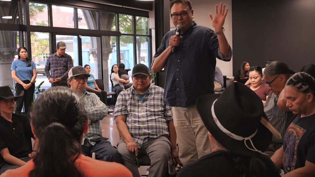 A man stands and speaks into a microphone, raising his hand, while seated individuals listen in a room with large windows and people standing in the background.