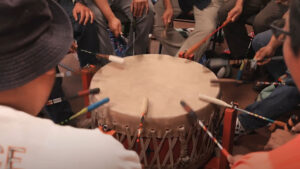 Several people sit around a large drum, each holding a drumstick and striking the drum in unison.