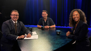 Three people sit at a round table with papers and mugs under studio lights, appearing ready for a discussion or interview.
