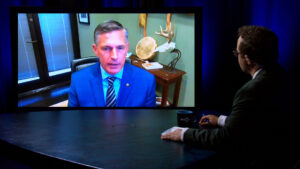 A man in a blue suit appears on a large video screen during a virtual interview, while another man in a suit sits at a table facing the screen.