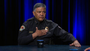 A police officer in uniform sits at a table with a blue mug, gesturing with one hand on his chest, against a dark blue curtain background.