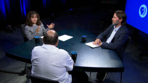 Three people sit around a table having a discussion in a studio setting, with papers and mugs on the table and a PBS logo visible on a screen in the background.