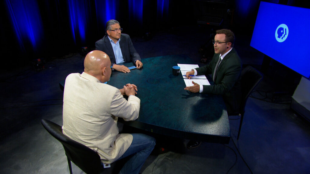 Three men sit around a round table in a TV studio, engaged in discussion. One man holds papers, while the other two listen. Blue lighting and a PBS logo screen are in the background.
