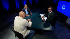 Three men sit around a round table in a TV studio, engaged in discussion. One man holds papers, while the other two listen. Blue lighting and a PBS logo screen are in the background.