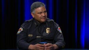 A police officer in uniform sits at a table holding a cup, with a dark blue and black background behind him.