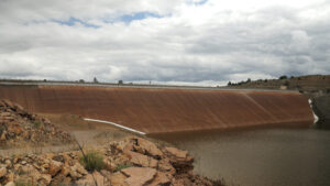 A large concrete dam with a sloped face holds back water under a cloudy sky, with rocky terrain in the foreground.