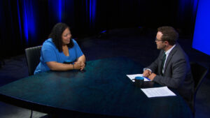Two people sit across from each other at a round table in a studio, engaged in conversation with papers and coffee mugs in front of them.