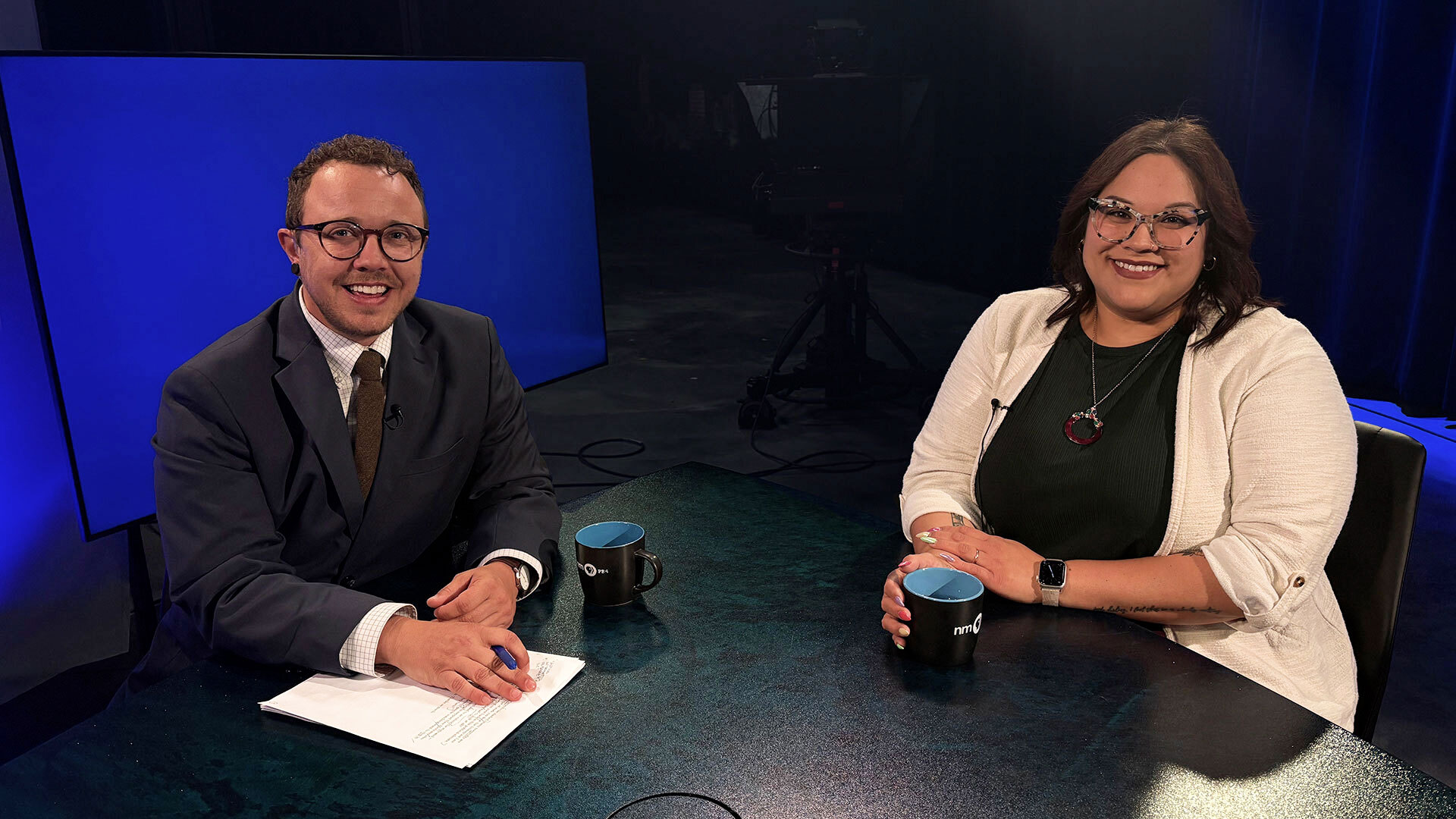 Two people sit at a table in a studio, facing the camera. Both are smiling, with mugs in front of them and papers on the table. A large blue screen is in the background.