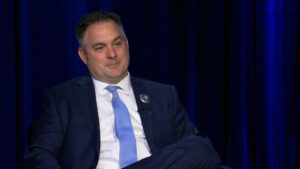 A man in a navy blue suit and light blue tie sits with his legs crossed against a dark blue curtain backdrop, looking slightly to the side.