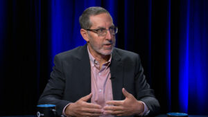 A man wearing glasses and a suit jacket gestures with his hands while speaking during an interview in a studio with blue lighting.