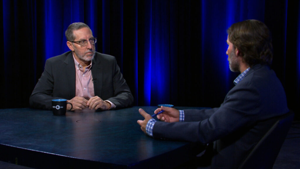 Two men in suits sit across from each other at a round table, having a conversation in a dimly lit studio with blue lights in the background.
