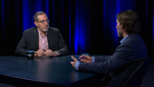 Two men in suits sit across from each other at a round table, having a conversation in a dimly lit studio with blue lights in the background.