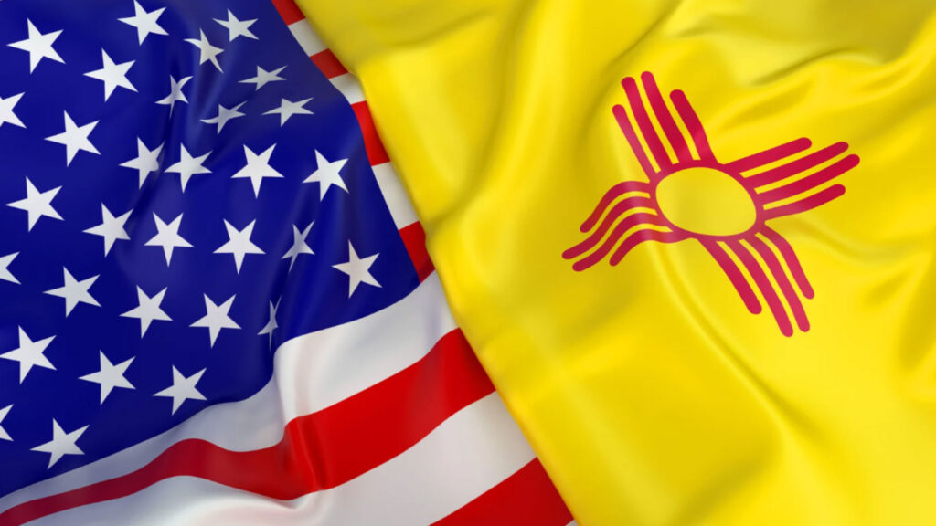 The United States flag and the New Mexico state flag are shown side by side, both flags appear to be gently waving.