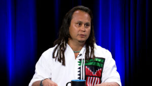 A man with long hair and dreadlocks, wearing a white shirt with colorful geometric patterns, sits at a table with a blue cup against a dark blue curtain background.