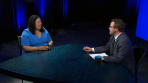 A woman and a man sit across from each other at a table in a studio, engaged in conversation. The man holds a pen and paper, and both have mugs. Blue lighting illuminates the scene.