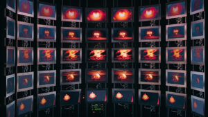 Multiple monitors arranged in a semicircle display repeated images of a nuclear explosion with a red-orange mushroom cloud against a dark background.