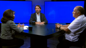 Three people sit around a black table having a discussion in a studio with blue screens in the background.