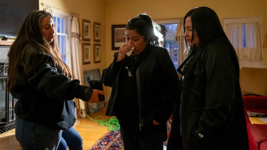 Three women stand in a living room, one showing something on her phone to the others; two women appear emotional or concerned.