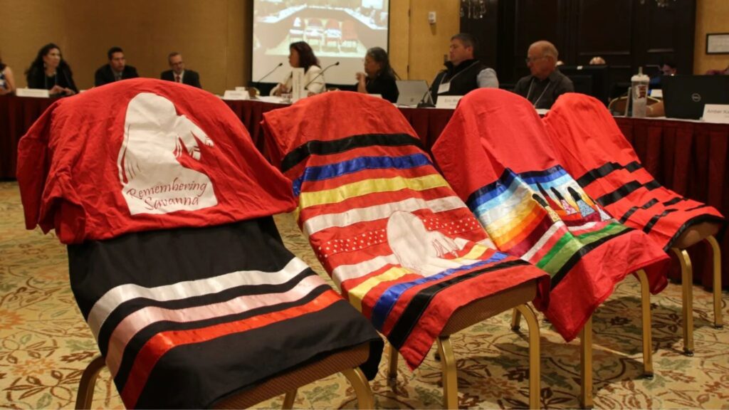 Four chairs draped with colorful blankets featuring Indigenous designs and the text "Remembering Savanna" at a conference panel discussion.