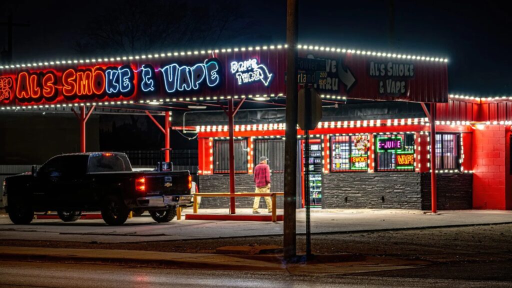 A person stands outside a brightly lit smoke and vape shop at night, with a black pickup truck parked nearby.