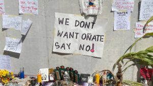 A protest altar with photos, candles, and signs, centered around a large banner that reads "WE DONT WANT OÑATE SAY NO!" taped to a concrete wall.