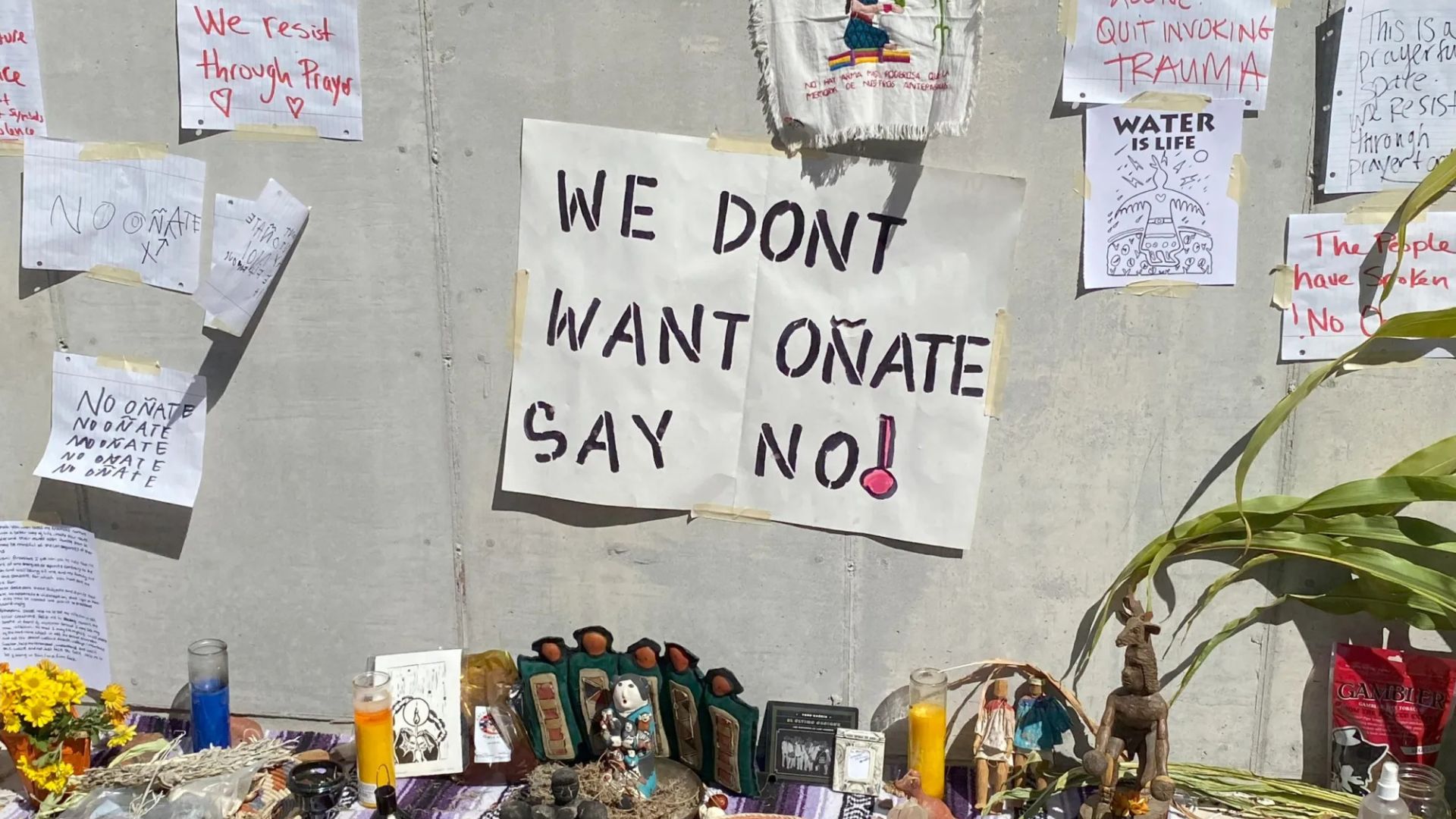A protest altar with photos, candles, and signs, centered around a large banner that reads "WE DONT WANT OÑATE SAY NO!" taped to a concrete wall.