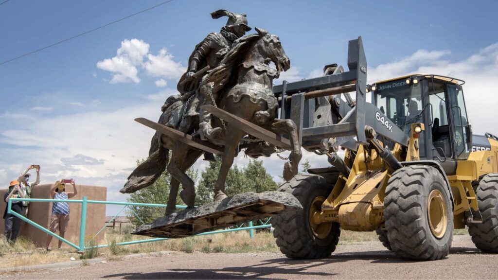 A front loader lifts a large bronze equestrian statue while two people observe and take photos in an outdoor setting.