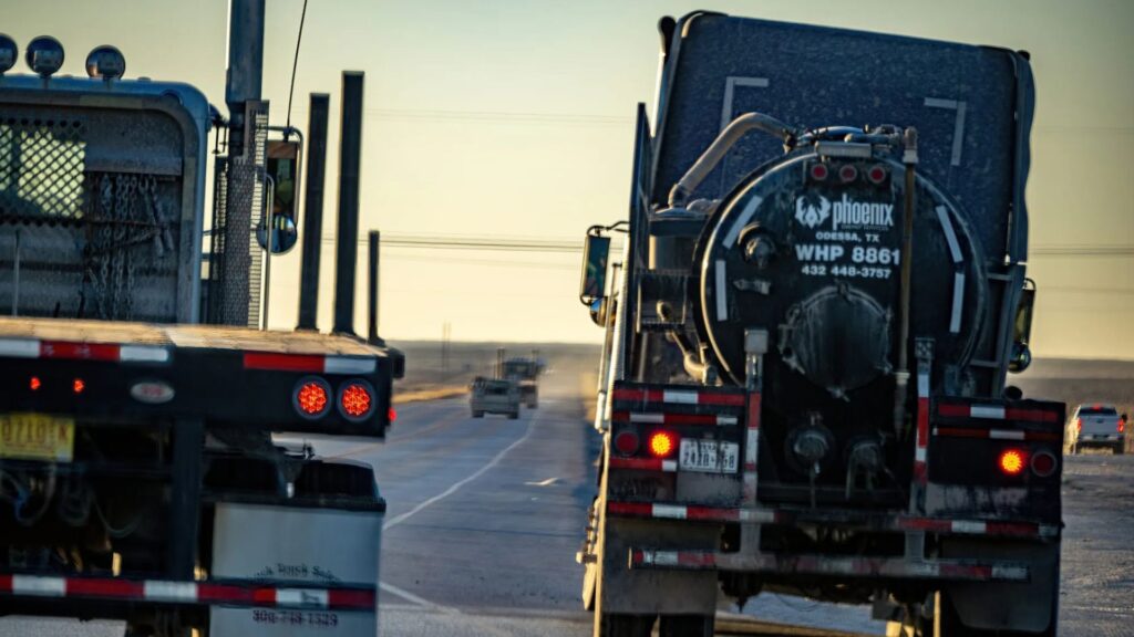 Two large trucks drive side by side on a rural highway under a clear sky, with another vehicle visible in the distance.