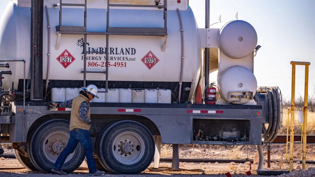 A worker in a hard hat walks beside a Timberland Energy Services tanker truck parked on a dirt lot.
