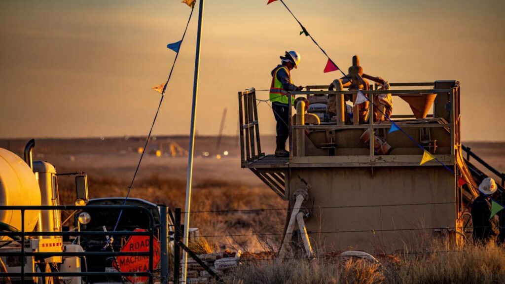 Workers in safety gear operate machinery on a raised platform in a desert area at sunset, with colorful pennant flags and vehicles nearby.