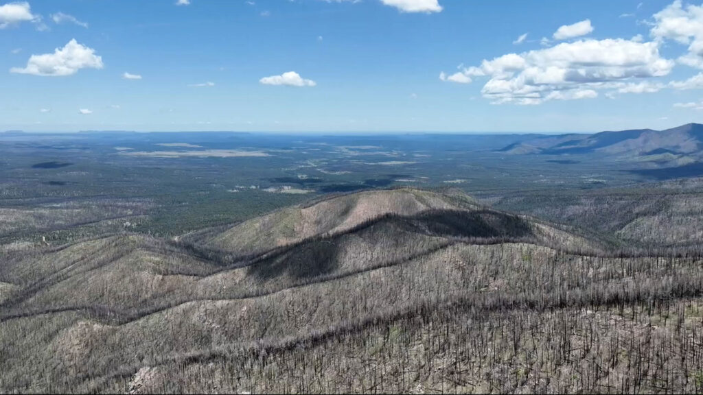 Aerial view of a mountain landscape with forested hills, some areas showing signs of previous wildfire damage, under a partly cloudy blue sky.